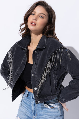 A light-wash denim jacket with a cropped length and long sleeves.  The jacket has a classic collar, two front pockets with button closures, and a button-down front.  The entire jacket is decorated with sparkling rhinestone fringe.